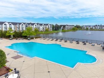 A large swimming pool with chaise lounge chairs next to a lake with apartment buildings in the